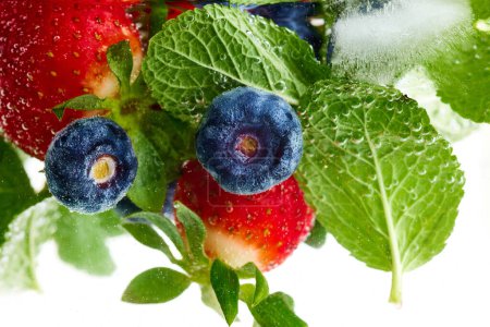 Nahaufnahme von frischer grüner Minze, Erdbeeren und Blaubeeren in Wasser mit Eis. Textur des kühlenden süßen Sommergetränks mit Makroblasen an der Glaswand. Konzept von Speisen und Getränken, Ernährung, Frische