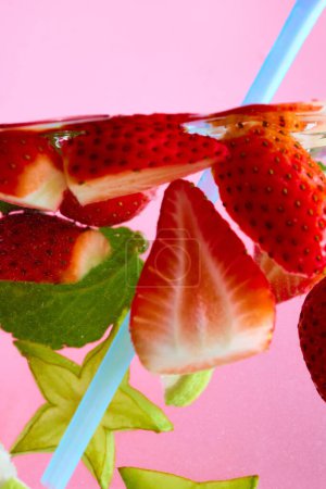 Aufgeschnittene Erdbeeren und grüne Blätter, die im Wasser schwimmen, mit einem hellblauen Strohhalm vor rosa Hintergrund. Textur des kühlenden süßen Sommergetränks. Konzept der Speisen und Getränke, Ernährung, Frische. Anzeige