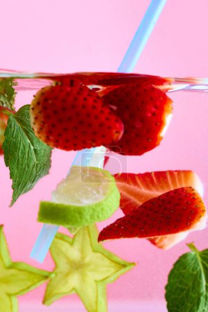 Erdbeeren in Scheiben geschnitten und grüne Minze im Wasser schwimmend mit hellblauem Stroh vor rosa Hintergrund. Textur des kühlenden süßen Sommergetränks. Konzept von Speisen und Getränken, Ernährung, Frische