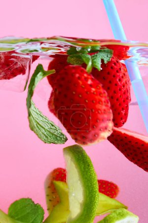 Geschnittene Erdbeeren, lindgrüne und grüne Blätter, die im Wasser schwimmen, mit hellblauem Stroh vor rosa Hintergrund. Textur des kühlenden süßen Sommergetränks. Konzept von Speisen und Getränken, Ernährung, Frische