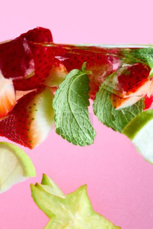 In Scheiben geschnittene Erdbeeren, Limette, Zitrone, Sternfrüchte und grüne Minze schwimmen im Wasser mit hellblauem Stroh vor rosa Hintergrund. Textur des kühlenden süßen Sommergetränks. Konzept von Essen, Trinken, Ernährung