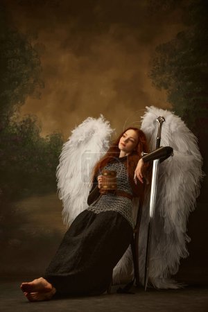 Redhead woman looks as angel in chainmail with sword, with large white wings and golden halo drinking beer against vintage studio background. Concept of historical and modern times, beauty and fashion