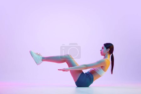 Téléchargez les photos : Jeune femme sportive assise et entraînement des muscles de l'abdomen en pose statique au néon sur fond de studio dégradé. Concept de sport et de loisirs, mouvement, autosoin, action, énergie. Publicité - en image libre de droit