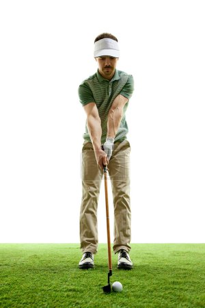 Focused golfer at start of powerful drive against white studio background. Skilled golf player stands on green grass with golf club. Concept of professional sport, luxury games, active lifestyle. Ad
