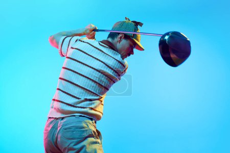 Rear view of man completing golf swing n mid follow-through with focus on club in neon light against gradient blue background. Concept of professional sport, luxury games, active lifestyle, action. Ad
