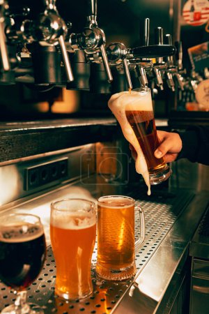 Foto von Barkeeper, der an der Bar mit mehreren Zapfsäulen leichtes, schäumendes Bier aus dem Zapfhahn in ein Pint-Glas gießt. Dazu Craft Beer vom Fass. Alkoholkonsum, Nachtleben, Feste, Oktoberfest.