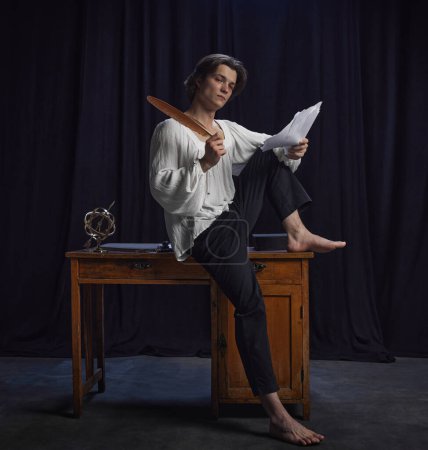 Young man, poet in old-fashion costume sitting on vintage desk and writing poem with feather against dark studio background. Concept of comparisons of eras, renaissance, friendship, inspiration.