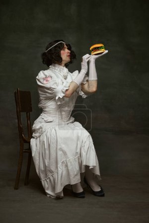 Photo for Young man in old-fashion dress, as woman holds huge delicious hamburger against vintage studio background. Concept of food and drink, self-expression, comparisons of eras, freedom, human rights. - Royalty Free Image