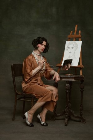 Photo for Man in silky dress with pearls seated by easel, reflecting on mirror behind and self-portrait against vintage studio background. Concept of self-expression, comparisons of eras, freedom, human rights. - Royalty Free Image