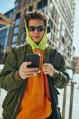 Photo for Young man in stylish sunglasses and headphones and bright casual attire stands confidently outside and using phone. Concept of street fashion and urban style, modern lifestyle, gen Z, self-expression. - Royalty Free Image