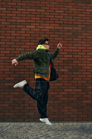 Photo for Trendy dressed young guy in sporting jacket and sunglasses jumping in motion against brick backdrop. Concept of street fashion and urban style, modern lifestyle, gen Z, self-expression. - Royalty Free Image