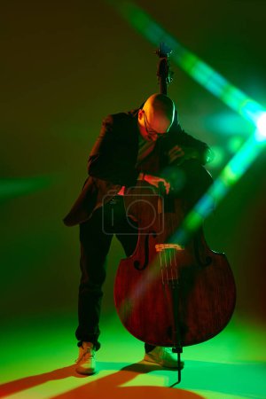 Photo for Portrait of bald musician in glasses performing on double bass in red-green neon light against gradient studio background. Concept of music and art, hobby, concerts and festivals, modern culture. Ad - Royalty Free Image