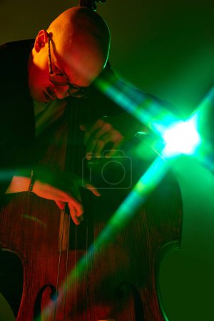 Téléchargez les photos : Musicien talentueux chauve à lunettes jouant du violoncelle en néon rouge-vert sur fond de studio dégradé. Concept de musique et d'art, passe-temps, concerts et festivals, culture moderne. Publicité - en image libre de droit