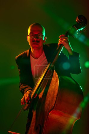 Photo for Stylish, contemporary bassist in glasses performs in red-green neon stage light against gradient studio background. Concept of music and art, hobby, concerts and festivals, modern culture. Ad - Royalty Free Image