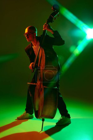 Photo for Double bass player performing contemporary jazz composition in red-green neon light against gradient studio background. Concept of music and art, hobby, concerts and festivals, modern culture. Ad - Royalty Free Image