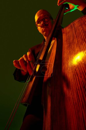 Photo for Artistic, talented young bass player performing jazz composition in red-green neon light against gradient studio background. Concept of music and art, hobby, concerts and festivals, modern culture. Ad - Royalty Free Image