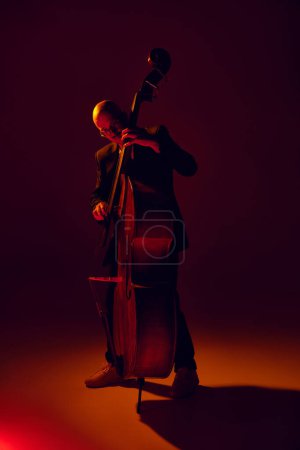 Glatzköpfiger, talentierter Bassist, der Jazzkompositionen im rot-gelben Licht vor abfallendem Studiohintergrund performt. Konzept von Musik und Kunst, Hobby, Konzerte und Festivals, moderne Kultur. Anzeige