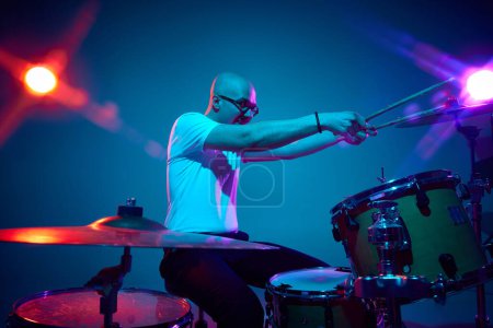 Photo for Artistic bald drummer in casual outfit with drumsticks in pink-purple stage lighting against gradient studio background. Concept of music and art, hobby, concerts and festivals, modern culture. Ad - Royalty Free Image