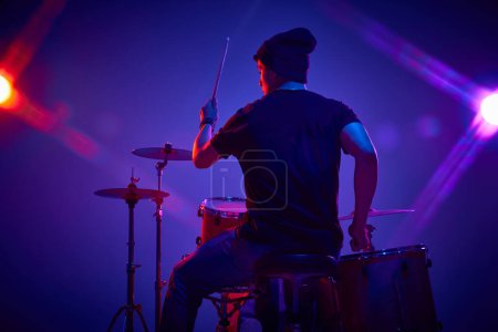 Photo for Rear view portrait of young artistic man playing drums in pink-purple stage lighting against gradient studio background. Concept of music and art, hobby, concerts and festivals, modern culture. Ad - Royalty Free Image