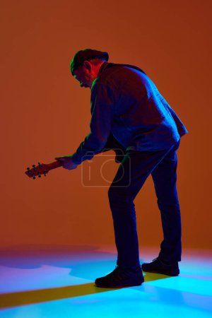 Photo for Rear view photo of mature musician in denim outfit playing guitar in colorful neon light against orange studio background. Concept of music, concerts and festivals, seniors in modern culture. Ad - Royalty Free Image