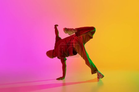 Dynamic photo of young, stylish dressed man breakdancer posing in neon light against gradient pink-yellow background. Concept of hobby, sport, creativity, fashion and style, motion, action. Ad