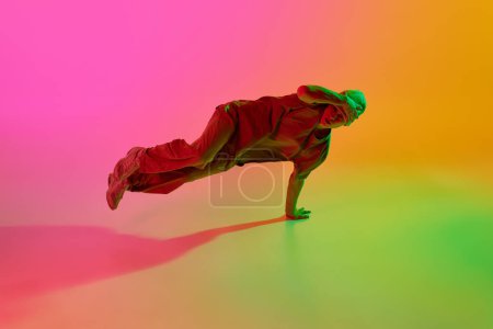 Young athlete man, dressed streetwear dancing hip-hop in motion in neon light against gradient pink-yellow background. Concept of art, hobby, sport, creativity, fashion and style, action. Ad