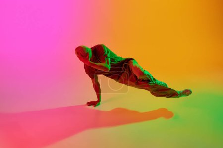 Young athlete man, dressed streetwear dancing freestyle moves in motion in neon light against gradient pink-yellow background. Concept of art, hobby, sport, creativity, fashion and style, action. Ad