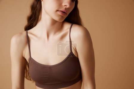 Close-up of young woman in brown sports bra against against sandy background. Well-kept neckline. Concept of beauty and health, skin care treatments, spa procedures. Ad
