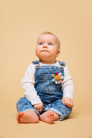 Photo for Baby with thoughtful expression looks upwards comfortably seated in soft denim overalls against warm beige background. Concept of beauty and fashion, childhood, motherhood, life, birth. Copy space. Ad - Royalty Free Image