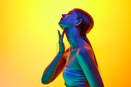 Photo for Side view photo of young sensual woman touching her neck in neon light against yellow gradient studio background. Concept of natural beauty, organic cosmetic, spa procedures, face-care. - Royalty Free Image