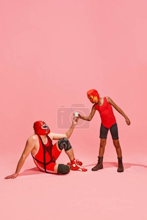Photo for Boy in superhero dressed red costume gave glass of milk to his older, bigger partner against pink studio background. Concept of pop art, generation difference, costume festivals, competitions. Ad - Royalty Free Image
