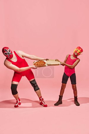 Dos luchadores, uno mayor y otro más joven, en trajes rojos luchando por pizza en caja sobre fondo de estudio rosa. Concepto de arte pop, diferencia de generación, festivales de disfraces, concursos. Anuncio
