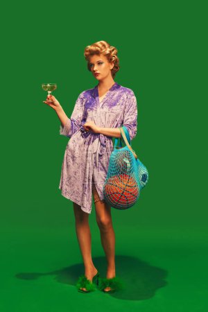 Photo for Young woman in silky bathrobe with hair curlers holding glass of cocktail sport balls against vibrant green background. Concept of sport, competition, active lifestyle, hobby, recreation. Ad - Royalty Free Image