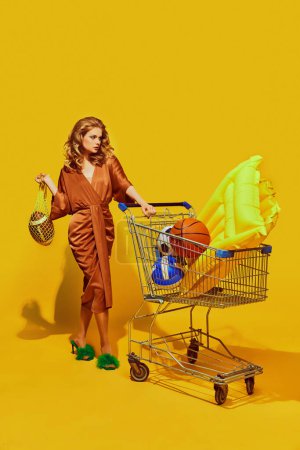 Frau in schickem braunem Kleid schiebt Wagen mit buntem Pool-Spielzeug und Sportgeräten vor gelbem Studiohintergrund. Konzept von Sport, aktiver Lebensweise, gesunder Ernährung, zeitgenössischer Kunst.