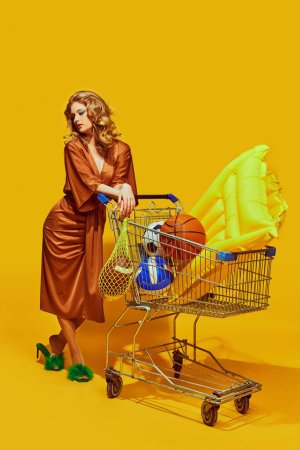 Elegante Dame hält Netztasche für Obst mit Sportball in der Hand und kauft Sommersachen vor gelbem Studiohintergrund ein. Konzept von Sport, aktivem Lebensstil und gesunder Ernährung, zeitgenössische Kunst.