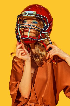 Photo for Portrait of woman in elegant attire wearing football helmet makes makeup against yellow studio background. Concept of woman in football, sport and fashion, style and beauty. Ad - Royalty Free Image