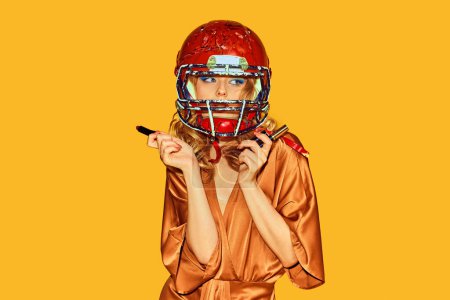 Photo for Beautiful woman in elegant attire wearing football helmet makes makeup against yellow studio background. Concept of woman in football, sport and fashion, style and beauty. Ad - Royalty Free Image
