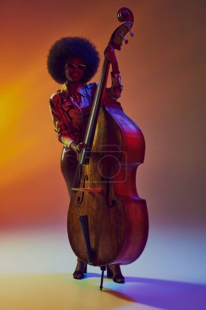 Stylish African-American woman, bass player with afro in neon light against gradient background. 70s funk vibe. Concept of art, music, hobby, classical music and modern lifestyle. Ad