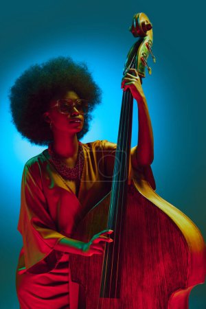 Photo for Vintage style African-American woman, jazz performer with glasses posing with double bass in neon light against gradient background. Concept of art, music, hobby, classical music and modern lifestyle. - Royalty Free Image