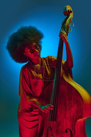 Retro dressed African-American woman, artist with curly hair posing with bass in neon light against gradient background. Concept of art, music, hobby, classical music and modern lifestyle. Ad
