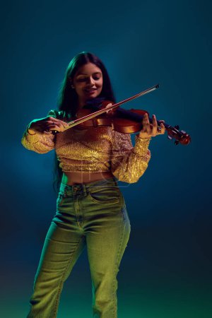 Artistic young Indian woman in glittery top enjoys playing violin in neon light against gradient background. Concept of art, music, hobby, classical music and modern lifestyle. Ad
