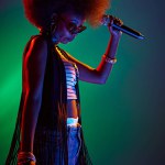 Afro-haired woman, talented vocalist in bohemian style performs in neon light against gradient background. Concept of art, music, hobby, classical music and modern lifestyle. Ad