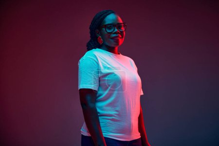Young attractive African-American woman dressed casual attire looking at camera in red neon light against gradient studio background. Concept of human emotions, beauty and fashion, style. Ad