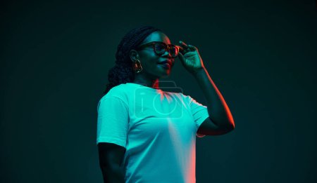 Seitenansicht Porträt einer lächelnden jungen Afroamerikanerin, die die Hand einer Brille in rotem Neonlicht vor den Studiohintergrund legt. Konzept menschlicher Emotionen, Schönheit und Mode, Stil.