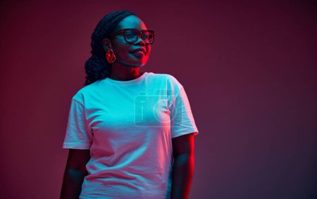 Selbstbewusste junge Afroamerikanerin in lässiger Kleidung und Brille, die im Neonlicht vor dem Studiohintergrund wegschaut. Konzept menschlicher Emotionen, Schönheit und Mode, Stil,