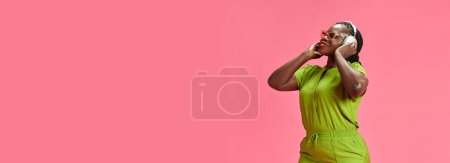 Banner. Joyful woman in vibrant green clothes listening music and dancing against pastel pink studio background with negative space. Concept of human emotions, fashion and beauty, trends.