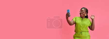 Banner. Young African-American woman waving hallo while looking at smartphone against pastel pink studio background. Copy space. Concept of human emotions, fashion and beauty, trends, connection. Ad
