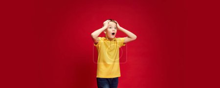 Banner. Surprised, shocked little boy put hands on head of excitement against red studio background with negative space. Concept of human emotions, childhood, education, fashion and style. Ad