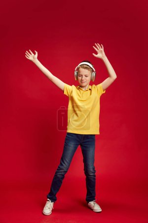 Portrait complet de l'adolescent, dansant tout en écoutant de la musique dans un casque sur fond de studio rouge. Petite danseuse. Concept d'émotions humaines, enfance, éducation, mode et style. Publicité