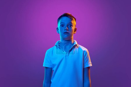 Portrait d'écolier en t-shirt décontracté à l'expression faciale choquée regardant la caméra en néon bleu clair sur fond dégradé violet. Concept d'enfance, d'éducation, de mode et de style. Publicité